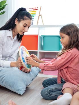 A woman with long, black hair up in a high ponytail holds up a game board for a young girl in a pink shirt.