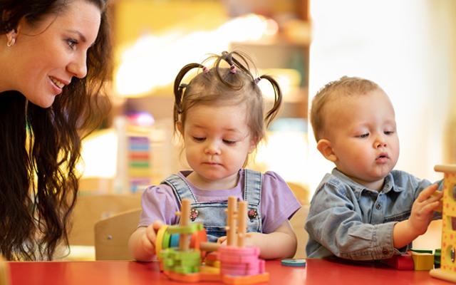 A Caucasian woman with wavy, dark brown hair watches over two toddlers who explore blocks.