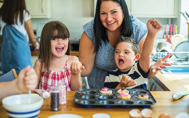 A smiling brunette woman helps small children with baking.