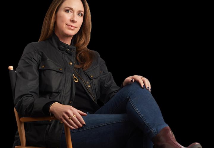 A white woman with straight brown hair sits cross-legged in a director's chair with a black background