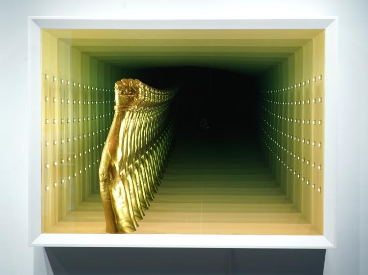 A metallic gold-painted sculpture of an arm with raised fist, situated within a white frame and surrounded by progressively smaller reflections.