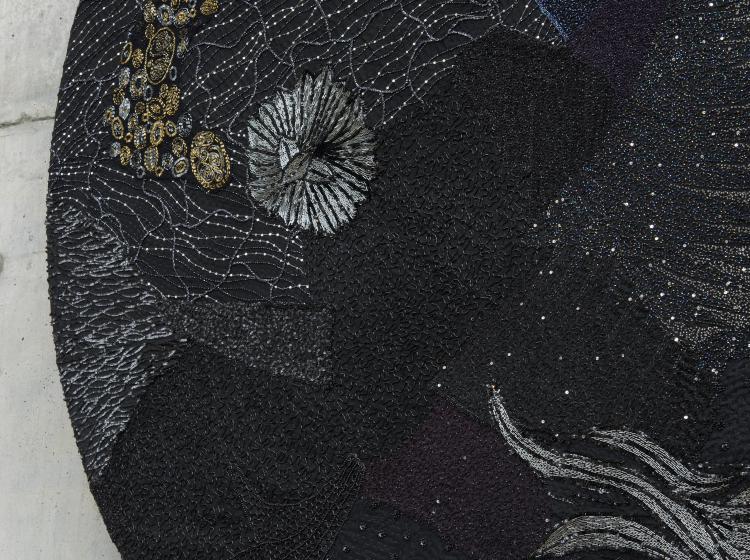 Close-up of a large, round multimedia artwork that is black and decorated in sequins and fabric, vaguely resembling stars and galaxies.