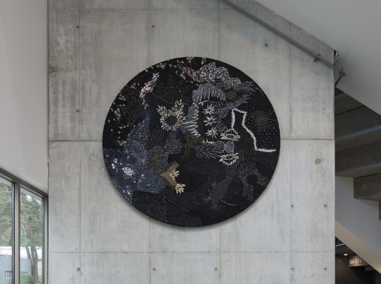 A large round multimedia artwork hangs on a wall in a well lit atrium area. The artwork is black and decorated in sequins and fabric, vaguely resembling stars and galaxies.