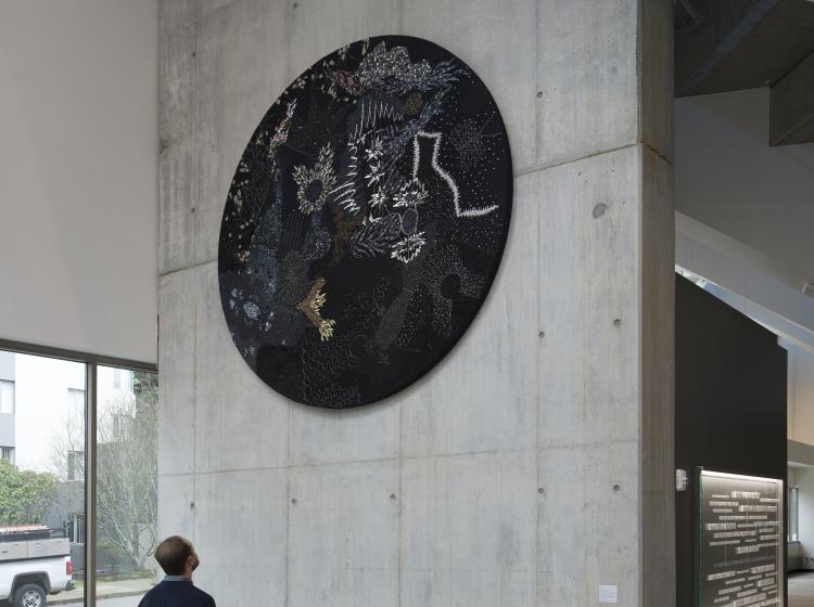 A large, round multimedia artwork that is black and decorated in sequins and fabric is hung high on a concrete wall, and a person stands looking at it.