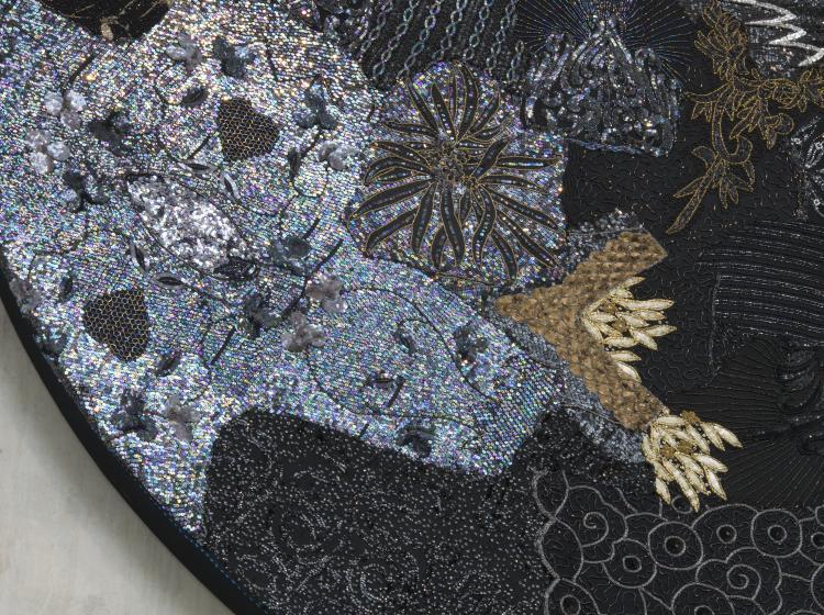 Close-up of a large, round multimedia artwork that is black and decorated in sequins and fabric, vaguely resembling stars and galaxies.