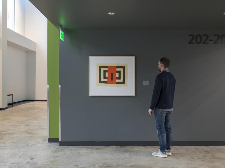 The artwork isframed on a dark grey wall in Peter Stott Center, with a person standing and viewing it.