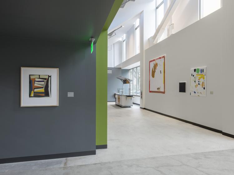The artwork is framed and hung on a dark grey wall in Peter Stott Center, at left. At right is a white hallway, with other artwork.