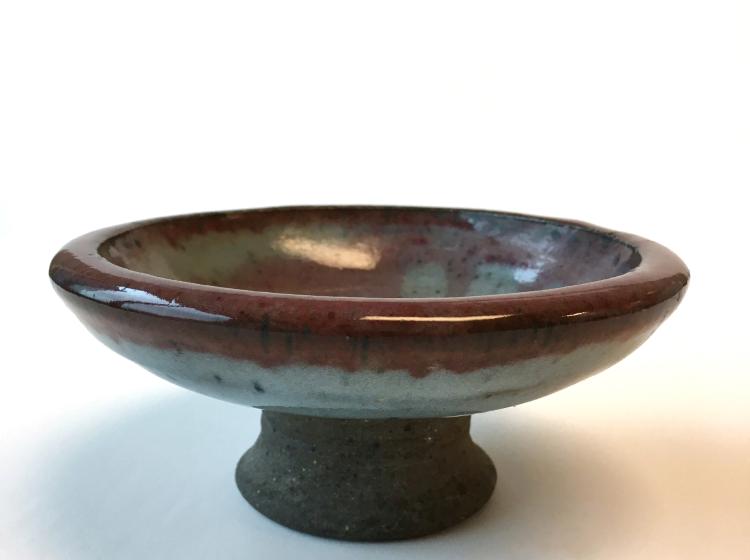 A ceramic dish against a white background. The dish has a small pedestal or foot, and is colored brown and celadon.