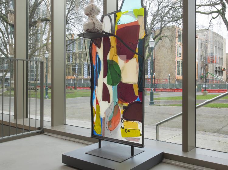 Angled view of a multi-colored abstract glass sculpture with irregular edges, framed in steel. The sculpture sits on a concrete floor in front of large windows.