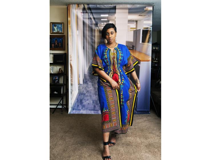 A young Black woman stands in front of a photo backdrop of a generic office suite. Her pose is confident, her gaze meeting the viewer. The backdrop is hung in a home environment, and a shelf of family photos can be seen at left.