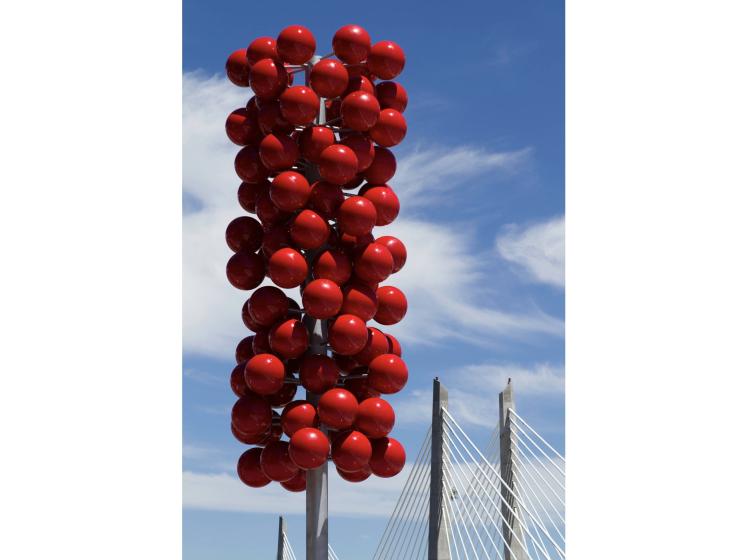 A large outdoor sculpture is seen on a sunny day, consisting of a steel pole with tightly spaced red orbs attached to the upper portion by short poles. Tilicum Crossing can be seen at right.