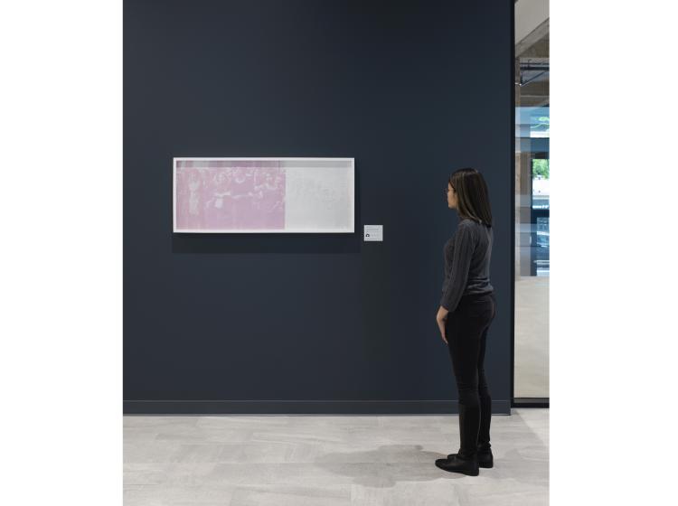 A framed photographic print is hung on a dark grey wall. Its colors are primarily white and pink, and the imagery is hard to make out. A person stands at right, looking at it.