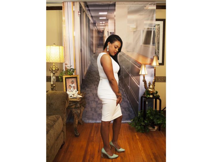 A young Black woman stands in front of a photo backdrop of a generic office suite. Her pose is confident, with gaze cast downward. The backdrop is hung in a home, with hardwood floor, a couch, plants and family photos.