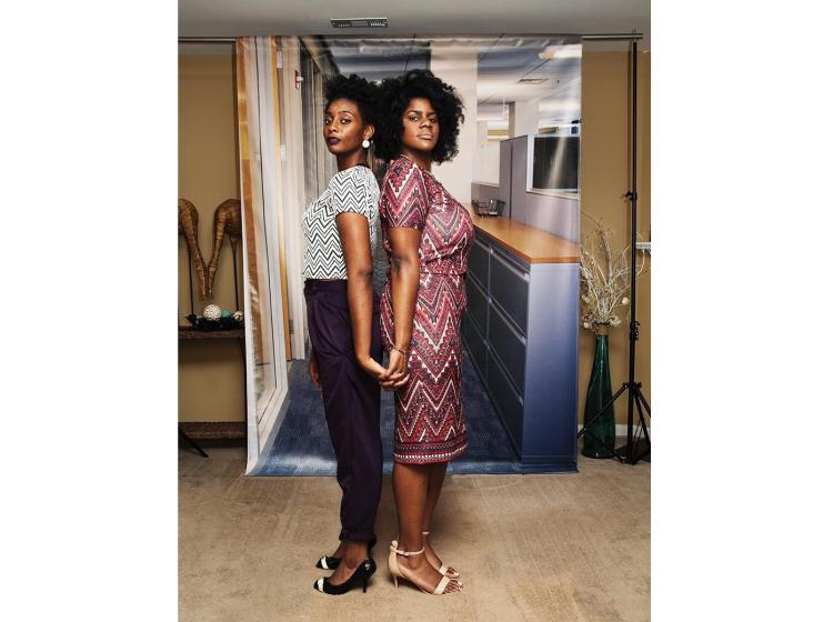 Two young Black women stand back-to-back in front of a photo backdrop of a generic office suite. The backdrop is hung in a home environment, and home decor can be seen in the background, such as a vase with plants.