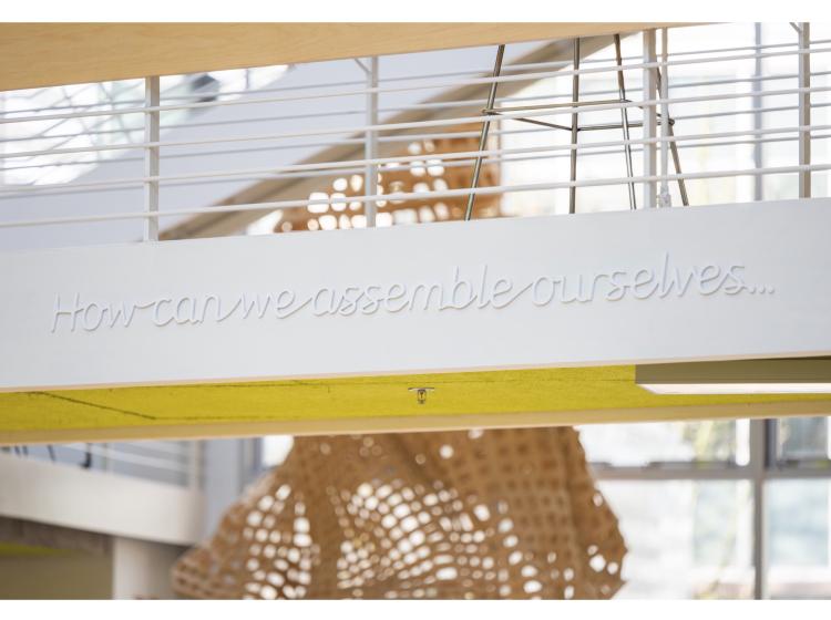 On the side of an elevated walkway in the Karl Miller Center atrium, white lettering in a raised italic font reads, “How can we assemble ourselves…”