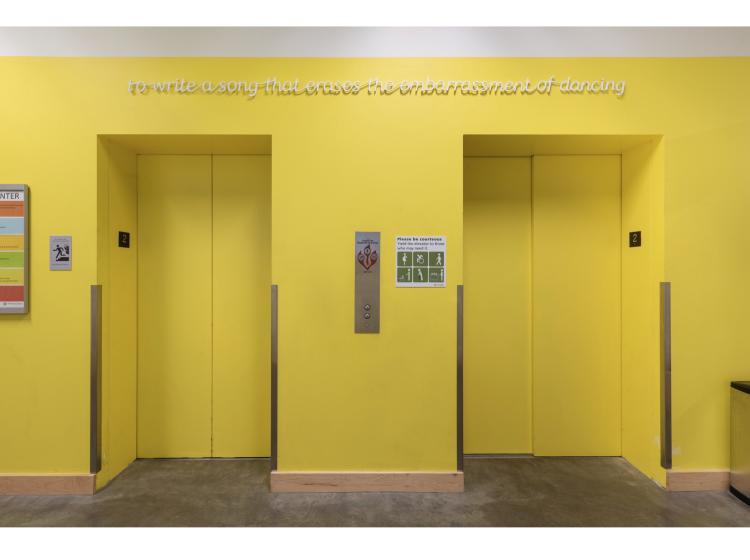 A wall and two elevator doors, all yellow. Above the elevators is raised white text reading, "to write a song that erases the embarrassment of dancing."