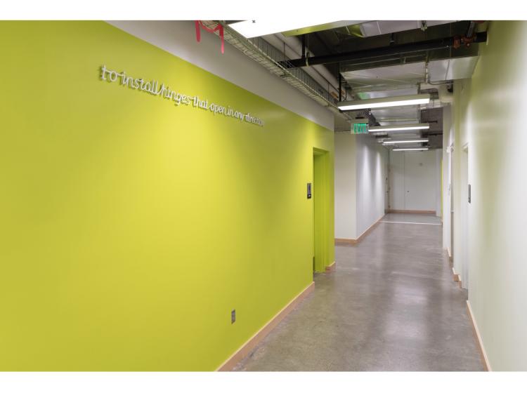 A hallway with the wall at left painted green and the wall at right painted white. On the green is a line of text in a raised white italic font, reading, “to install hinges that open in any direction.”
