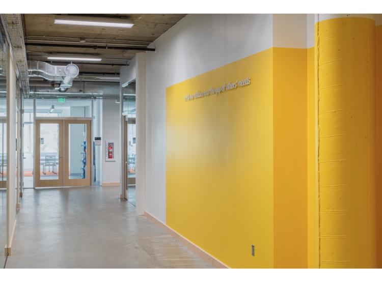 In a hallway, text appears on an orange wall in a raised white italic font. It reads, “to fit our abilities into the gaps of others’ needs.”