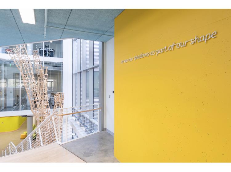 A bright golden orange wall at right. At left is the atrium of Karl Miller Center. On the orange wall is a line of text, in white italic font. It reads, “to see our shadows as part of our shape.”