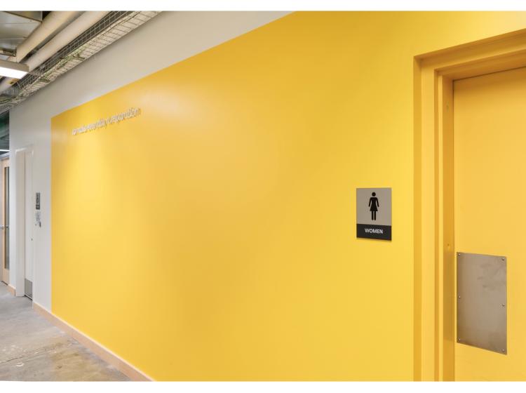 A bright golden-orange wall. At right, the door to a women’s restroom is seen. On the wall, a line of text appears in a raised white italic font. It reads, “to make every day a reparation.”