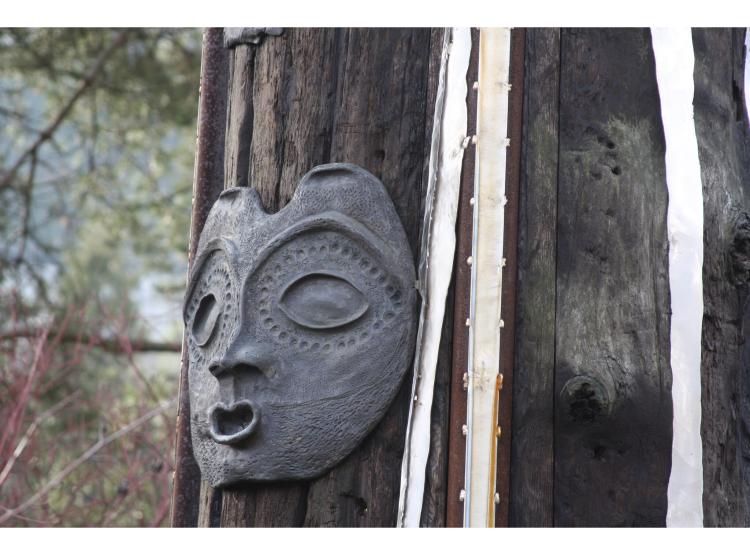 Close-up of a section of the pole. The wood is roughly textured and decorated with small sculptural elementsm. Featured here is an abstracted metal face.