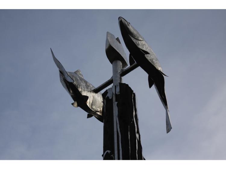 View from below of two large metal salmon at the top of the pole. They are swimming in opposite directions, attached just above the top of the pole.