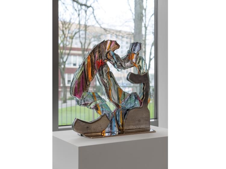 Multicolored glass abstract sculpture on a metal base, seen from a slight angle. The shape of the sculpture loosely resembles a pretzel. In the background, through a window, Shattuck Hall can be seen.