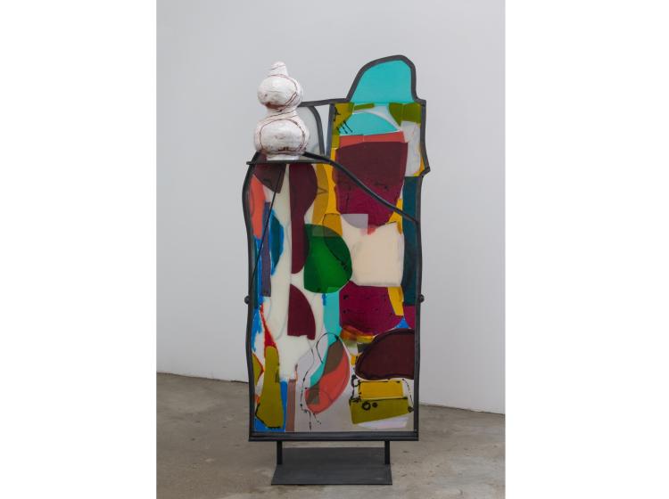 A multi-colored, fused glass abstract sculpture with irregular edges, framed in steel. The sculpture sits on a concrete floor in front of a white wall.