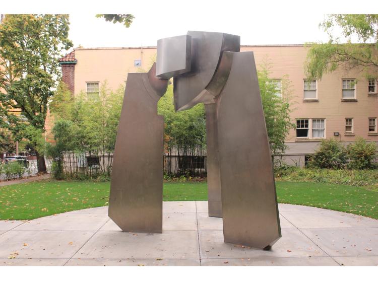 A large abstract steel sculpture on a round granite base surrounded by grass, buildings and trees. The sculpture has three “legs” meeting at the top in a joined area with a round hole through it.