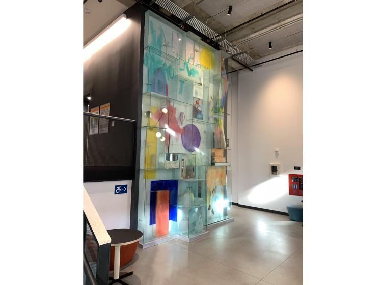 The entire artwork is seen from across the room. It is a floor-to-ceiling installation of glass geometric sections with various colorful elements and objects inside.
