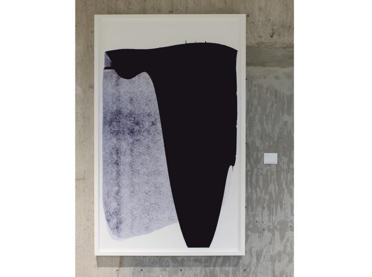 A large print with dark abstract shapes is seen framed on a concrete wall.
