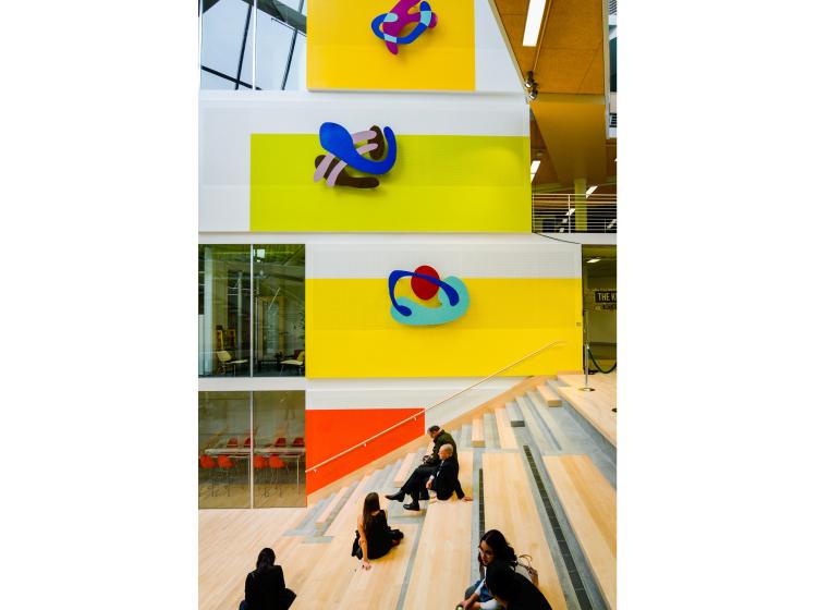 The artwork, composed of brightly colored abstract shapes against large colorful rectangles, is seen in the Karl Miller Center atrium, and in the foreground are wooden steps with students seated on them.