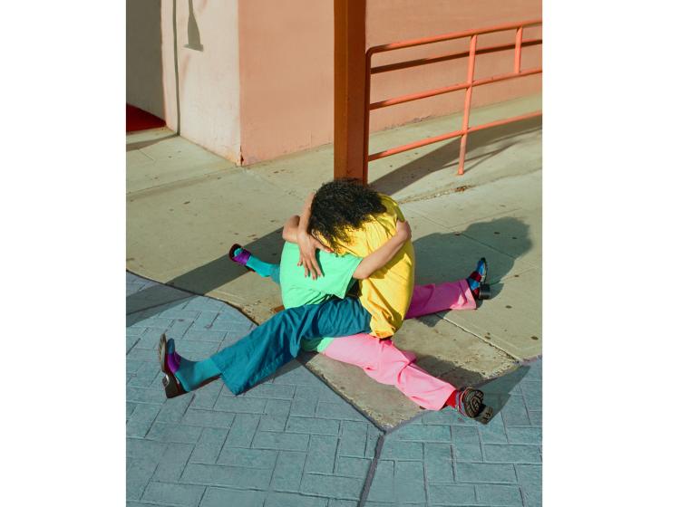 Two figures sit on a sidewalk corner embracing, with their legs splayed out to the sides, so that all four legs form an X shape. They are wearing vivid green, yellow, blue and pink clothing.