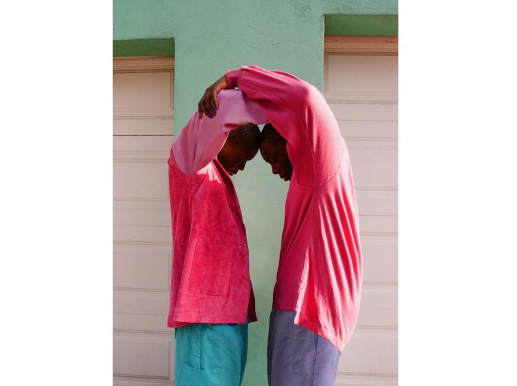 Vividly colored photo of two figures standing face to face, arms entwined above their heads so they form an arc shape. They both wear brilliant pink, in front of a green wall
