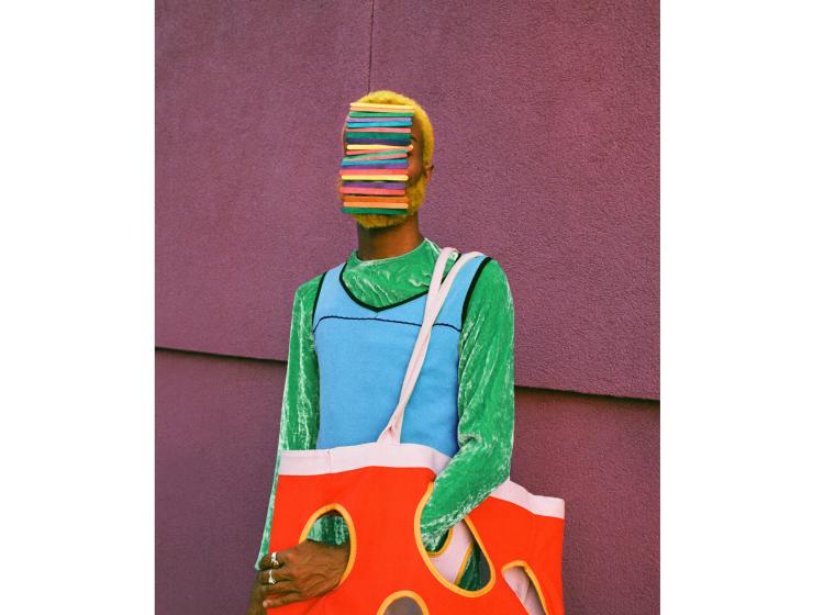 Vividly colored photo of a figure standing in front of a purple wall, wearing bright green and blue, holding a red tote bag with circular holes in it, and with multicolored popsicle sticks obscuring their face