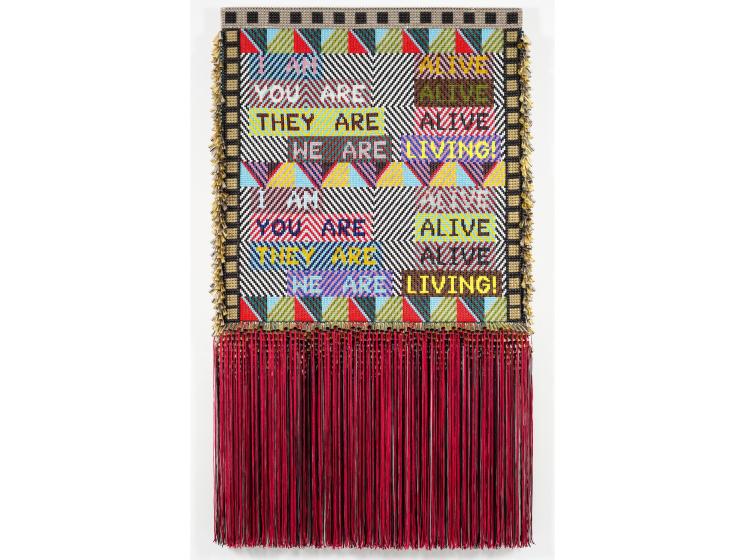 Large wall hanging, consisting of beadwork, stud panels, fringe, and jingles. The beaded text reads: "I AM ALIVE/YOU ARE ALIVE/THEY ARE ALIVE/WE ARE LIVING!" repeated twice in varying colors.