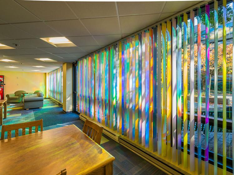 Interior of Blumel Hall's first floor resident lounge. In the long floor to ceiling windows, the blinds are covered in multicolored, reflective material, casting colored reflections of sunlight into the space