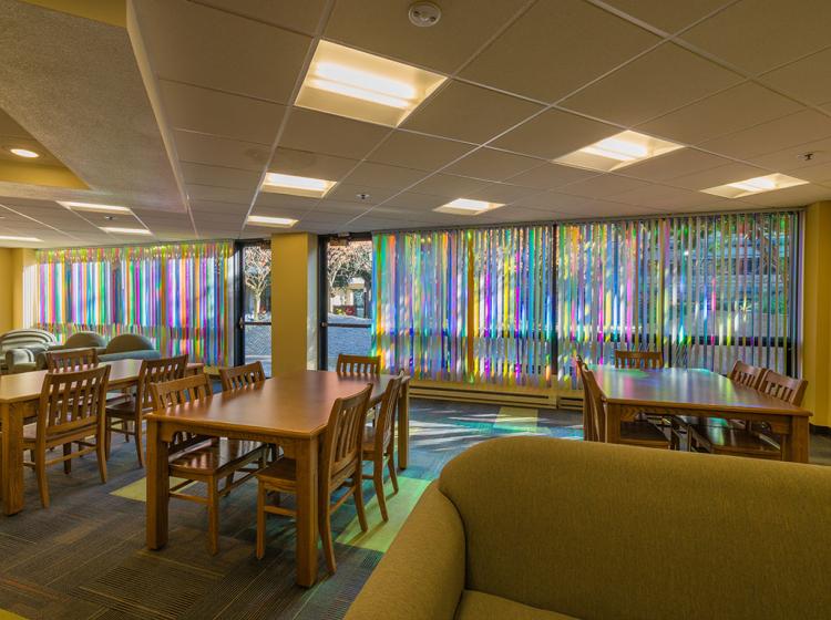 Wide shot of the student lounge with tables and chairs, with the multicolored reflective blinds in background