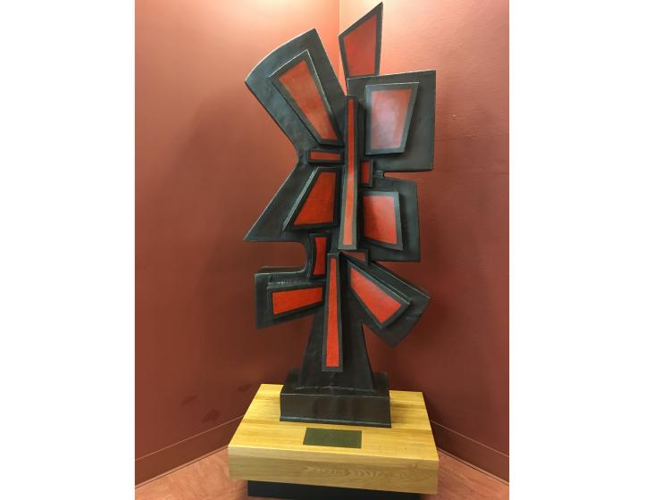 Abstract sculpture with variously shaped geometric forms jutting out at different angles, in black and red, on a wooden base