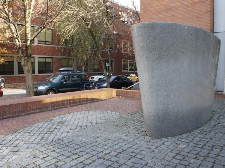 A large, abstract granite sculpture sits on the Urban Plaza. It is grey, shaped like a cross section of an inverted cone, and has a rough top surface and smooth sides.