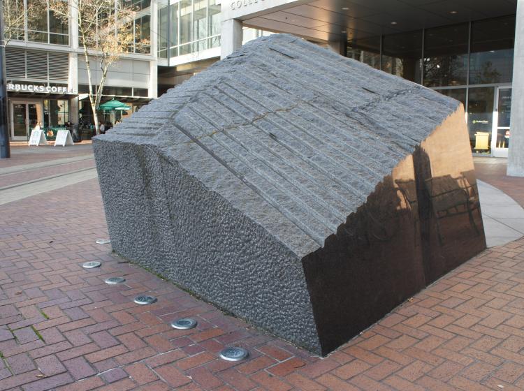 A large, abstract granite sculpture sits on the Urban Plaza, in front of the Urban Center. It is dark grey, roughly trapezoid shaped, with a ridged top surface and shiny smooth sides.