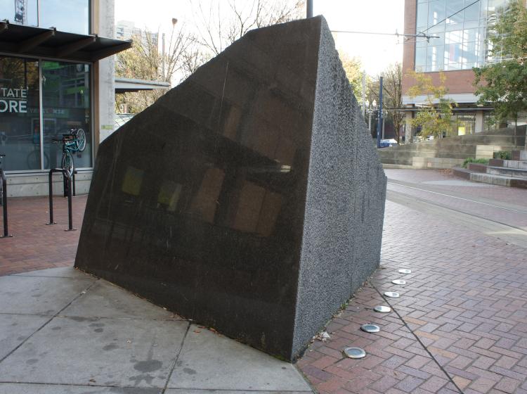 A large, abstract granite sculpture sits on the Urban Plaza, in front of the Urban Center. It is dark grey, roughly trapezoid shaped, with a smooth shiny side facing the viewer and another visible side that is rough in texture.