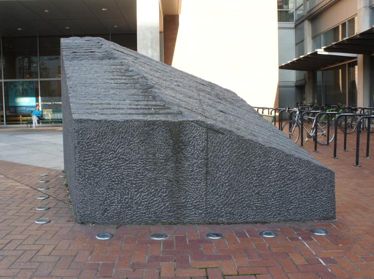 A large, abstract granite sculpture sits on the Urban Plaza, in front of the Urban Center. It is dark grey, roughly trapezoid shaped. Its sloped top surface is ridged, and the side facing the viewer has a rough texture.
