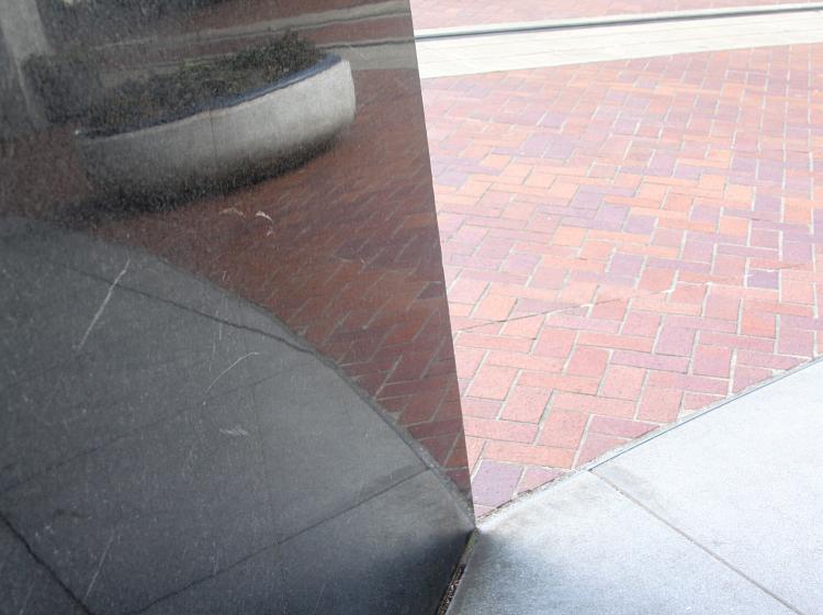Close-up shot of the lower portion of the darker sculpture. The brick plaza surface is seen at right, and the sculpture is at left, with the brick surface reflecting on it
