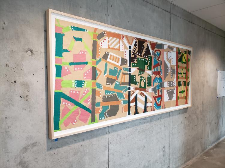 The artwork is seen framed and hung on a concrete wall, viewed at an angle from the left side