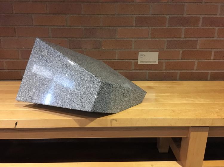 A small, abstract granite sculpture is seen on an interior wooden bench in front of a brick wall. The sculpture is roughly trapezoid shaped
