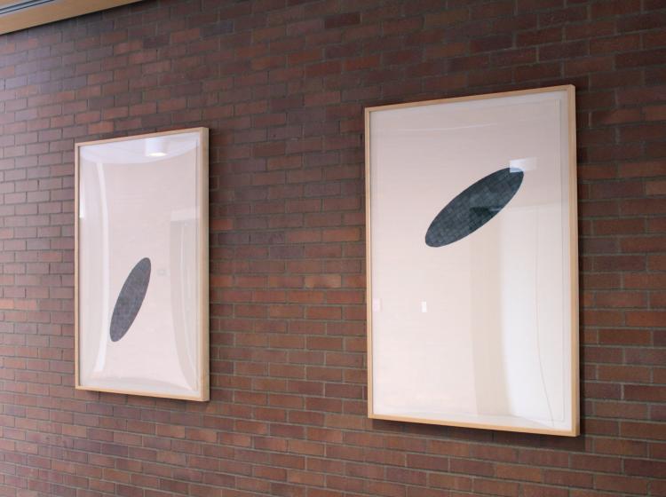 Two framed prints are seen on an interior brick wall. Each print has an abstract elliptical shape in black on white