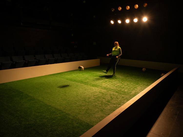 Cast member of PSU Theater's "The Wolves" dressed in soccer costumes, kicking a ball on astroturf in dramatic lighting.