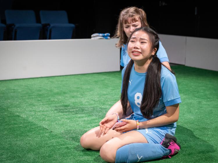 Cast of PSU Theater's "The Wolves" dressed in soccer costumes. One looking very upset, the other comforting her.