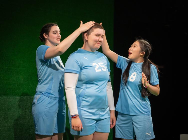 Cast of PSU Theater's "The Wolves" dressed in soccer costumes. Two players rubbing the buzzed hair of the other player.